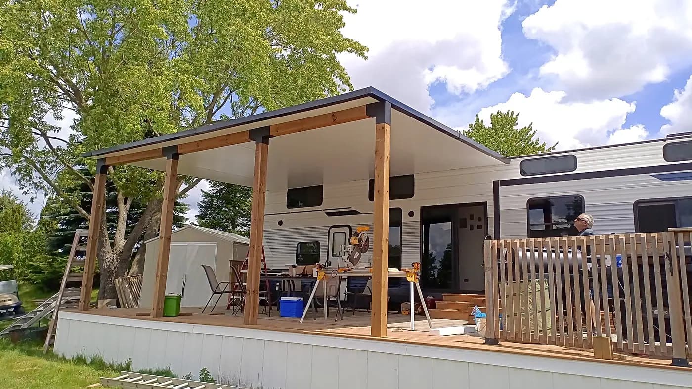 Large covered wooden deck with a pitched roof on posts, alongside a trailer
