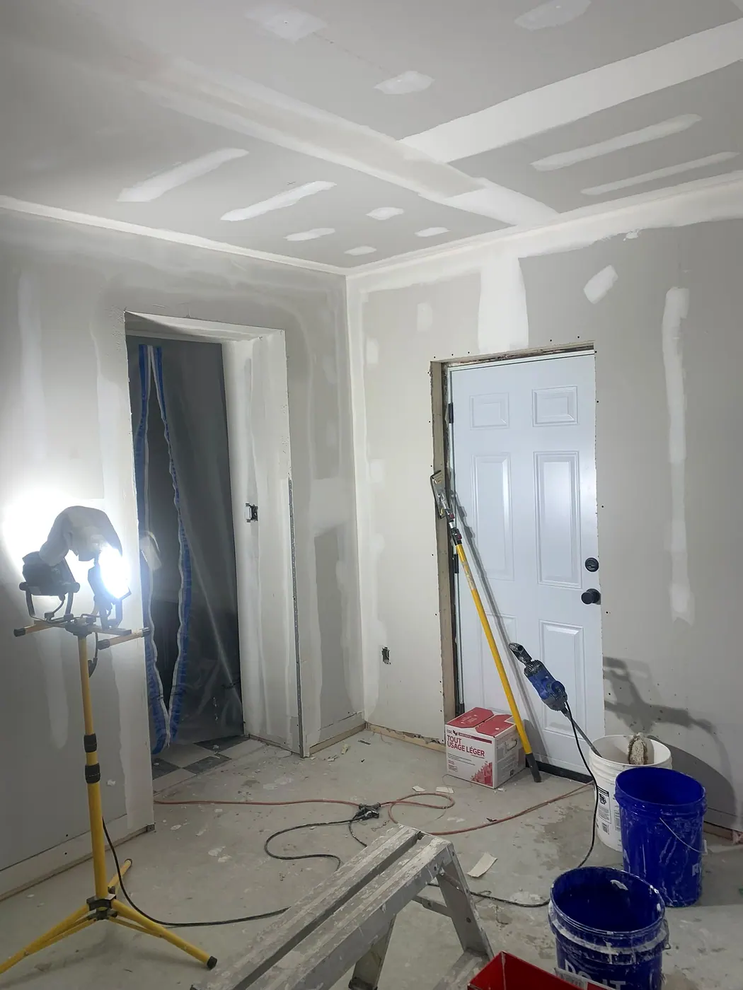 Hallway with drywall being taped around two door openings, lit by a work light